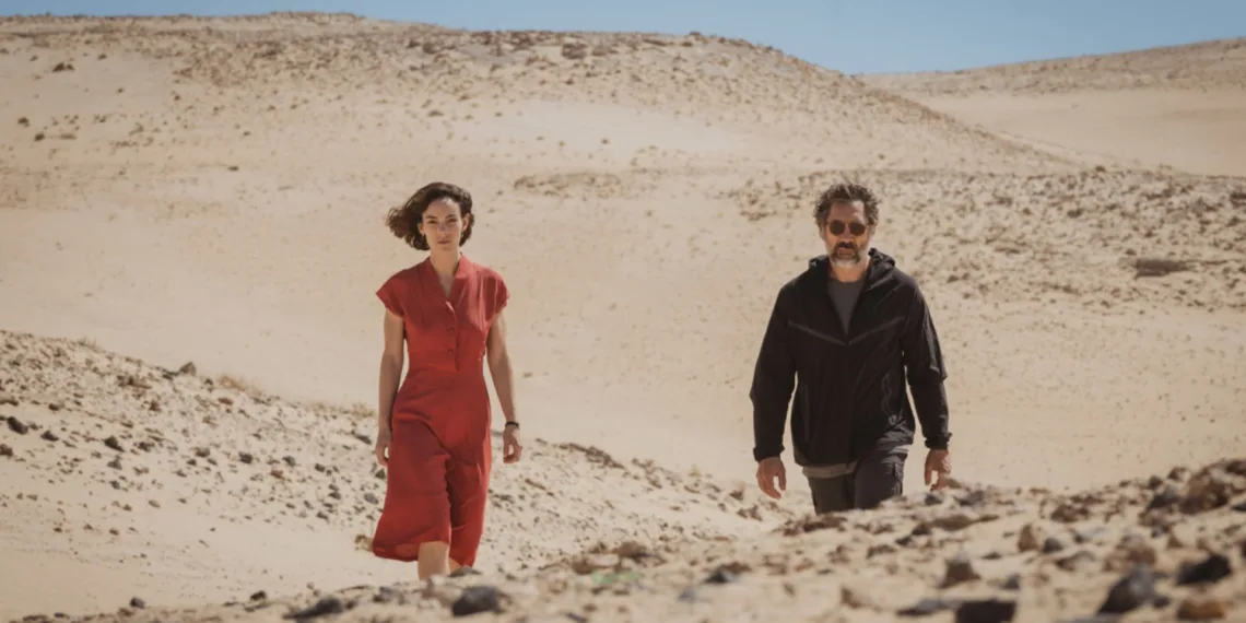 Rodrigo Sorogoyen’s cinema often studies the violence that gathers when bodies, memories, and egos are forced into close range. In The Beloved, that pressure hardens under the sun-blasted dryness of Fuerteventura. Esteban Martínez, a celebrated Oscar-winning Spanish filmmaker, returns to his birthplace after a long, profitable stay inside the American studio system. He arrives speaking the language of renewal. He offers an apparent peace gesture to Emilia, his estranged daughter, an actress still searching for the role that might give her public shape. The gift is a principal part in Desert, his historical epic about the 1930s Sahrawi uprising against Spanish colonial oppression. The scorched volcanic island becomes a moral furnace. Sorogoyen turns the film set into a psychological enclosure where artistic power presses against the jagged remains of family damage. Before the cameras begin their ritual work, the production already resembles a clinical chamber for old ghosts, lit by the pitiless stare of a director who mistakes control for truth. The Architecture of Fractured Memory The opening movement establishes its sickness through a static twenty-minute restaurant sequence in Madrid. Sorogoyen lets estrangement speak through posture, hesitation, appetite, and silence. Emilia reaches for red wine and beer, building a small chemical shelter around herself. Esteban sips sparkling water with theatrical restraint, projecting the serenity of a man who has rehearsed sobriety as a public image. Their past has the shape of a wound that refuses a single witness. Emilia remembers a childhood trauma, a screening of Kill Bill Volume 2 marked by her father’s violent drunken abuse. Esteban receives the memory with calm denial and recasts his own cruelty as a harmless invention. Javier Bardem gives him a predatory magnetism, a quiet menace that covers the history of his volatility like fresh paint over damp rot. His aging auteur treats repentance as material to be staged, blocked, revised, and owned. Victoria Luengo meets that force with extraordinary defensive vulnerability. Her face flickers from tentative hope to self-loathing, then to anger that seems to arrive years late, carrying the sediment of neglect. The casting choice feels darkly solipsistic. Esteban appears to have built the entire desert production as an elaborate trap, using professional patronage to draw from his daughter the submission and forgiveness she has never granted him freely. The Autocracy of the Frame Inside the Desert production, the movie set becomes a sealed kingdom, a feudal organism where artistic creation grants permission for absolute rule. Sorogoyen reveals the brittleness of that authority during a pivotal country lunch sequence. Beneath a punishing morning sun, the actors sink into repeated errors and fail to deliver their lines with the demanded conviction. Esteban’s irritation hardens into icy sadism, and his rage fixes on their inability to eat a plate of fish stew with adequate appetite. The scene turns a creative exercise into psychological punishment, exposing the terror beneath his control. The supporting figures mark the limits of this poisoned space. Marina, the exhausted producer, tries to contain his outbursts, becoming a living bridge from an era of unchecked administrative power to one of modern accountability. The female cinematographer chooses exile from his rule and resigns once his behavior becomes impossible to bear. Her departure signals a major change in contemporary industry standards, where the old impunity of the tyrannical director now meets collective resistance. The film finds its sharpest moral wound here. Esteban directs an expensive work of anti-colonial art, lectures viewers on imperial dominance, and colonizes the psyches of his crew and family with the same spiritual violence he claims to condemn. Shifting Stocks and Fractured Truths Rodrigo Sorogoyen’s cinema often studies the violence that gathers when bodies, memories, and egos are forced into close range. In The Beloved, that pressure hardens under the sun-blasted dryness of Fuerteventura. Esteban Martínez, a celebrated Oscar-winning Spanish filmmaker, returns to his birthplace after a long, profitable stay inside the American studio system. He arrives speaking the language of renewal. He offers an apparent peace gesture to Emilia, his estranged daughter, an actress still searching for the role that might give her public shape. The gift is a principal part in Desert, his historical epic about the 1930s Sahrawi uprising against Spanish colonial oppression. The scorched volcanic island becomes a moral furnace. Sorogoyen turns the film set into a psychological enclosure where artistic power presses against the jagged remains of family damage. Before the cameras begin their ritual work, the production already resembles a clinical chamber for old ghosts, lit by the pitiless stare of a director who mistakes control for truth. The Architecture of Fractured Memory The opening movement establishes its sickness through a static twenty-minute restaurant sequence in Madrid. Sorogoyen lets estrangement speak through posture, hesitation, appetite, and silence. Emilia reaches for red wine and beer, building a small chemical shelter around herself. Esteban sips sparkling water with theatrical restraint, projecting the serenity of a man who has rehearsed sobriety as a public image. Their past has the shape of a wound that refuses a single witness. Emilia remembers a childhood trauma, a screening of Kill Bill Volume 2 marked by her father’s violent drunken abuse. Esteban receives the memory with calm denial and recasts his own cruelty as a harmless invention. Javier Bardem gives him a predatory magnetism, a quiet menace that covers the history of his volatility like fresh paint over damp rot. His aging auteur treats repentance as material to be staged, blocked, revised, and owned. Victoria Luengo meets that force with extraordinary defensive vulnerability. Her face flickers from tentative hope to self-loathing, then to anger that seems to arrive years late, carrying the sediment of neglect. The casting choice feels darkly solipsistic. Esteban appears to have built the entire desert production as an elaborate trap, using professional patronage to draw from his daughter the submission and forgiveness she has never granted him freely. The Autocracy of the Frame Inside the Desert production, the movie set becomes a sealed kingdom, a feudal organism where artistic creation grants permission for absolute rule. Sorogoyen reveals the brittleness of that authority during a pivotal country lunch sequence. Beneath a punishing morning sun, the actors sink into repeated errors and fail to deliver their lines with the demanded conviction. Esteban’s irritation hardens into icy sadism, and his rage fixes on their inability to eat a plate of fish stew with adequate appetite. The scene turns a creative exercise into psychological punishment, exposing the terror beneath his control. The supporting figures mark the limits of this poisoned space. Marina, the exhausted producer, tries to contain his outbursts, becoming a living bridge from an era of unchecked administrative power to one of modern accountability. The female cinematographer chooses exile from his rule and resigns once his behavior becomes impossible to bear. Her departure signals a major change in contemporary industry standards, where the old impunity of the tyrannical director now meets collective resistance. The film finds its sharpest moral wound here. Esteban directs an expensive work of anti-colonial art, lectures viewers on imperial dominance, and colonizes the psyches of his crew and family with the same spiritual violence he claims to condemn. Shifting Stocks and Fractured Truths Cinematographer Álex de Pablo gives this psychic dissonance an unstable visual language. The film moves through an erratic mutation of media, with scenes captured on 65mm, 35mm, 16mm, and 8mm celluloid stocks, along with cold digital footage and grainy video-assist textures. Abrupt shifts from monochrome landscapes to sudden color passages disturb any stable narrative ground. At times, one wonders if this aesthetic volatility reaches past its own necessity. Still, it reflects the atmosphere of a set ruled by impulse, making the viewer inhabit Emilia’s disorientation. Sorogoyen roots this formal experimentation in a fictional archive by inserting clips from Esteban’s 1990s debut film, Siroco, starring Emilia’s mother, Charo Vera. The texture carries the chill of ancestral abandonment, the old image returning like a debt never paid. The narrative invokes Liv Ullmann’s observation that proximity to a camera requires the removal of one’s mask. Under the technical pressure of the final scenes, that idea takes full shape. Tight framing breaks down Esteban’s ego and releases a buried reservoir of regret. The sound design extends the fragmentation through a dissonant score and fractured soundscapes, echoing the irreconcilable versions of domestic truth carried by the characters.