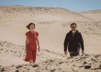Rodrigo Sorogoyen’s cinema often studies the violence that gathers when bodies, memories, and egos are forced into close range. In The Beloved, that pressure hardens under the sun-blasted dryness of Fuerteventura. Esteban Martínez, a celebrated Oscar-winning Spanish filmmaker, returns to his birthplace after a long, profitable stay inside the American studio system. He arrives speaking the language of renewal. He offers an apparent peace gesture to Emilia, his estranged daughter, an actress still searching for the role that might give her public shape. The gift is a principal part in Desert, his historical epic about the 1930s Sahrawi uprising against Spanish colonial oppression. The scorched volcanic island becomes a moral furnace. Sorogoyen turns the film set into a psychological enclosure where artistic power presses against the jagged remains of family damage. Before the cameras begin their ritual work, the production already resembles a clinical chamber for old ghosts, lit by the pitiless stare of a director who mistakes control for truth. The Architecture of Fractured Memory The opening movement establishes its sickness through a static twenty-minute restaurant sequence in Madrid. Sorogoyen lets estrangement speak through posture, hesitation, appetite, and silence. Emilia reaches for red wine and beer, building a small chemical shelter around herself. Esteban sips sparkling water with theatrical restraint, projecting the serenity of a man who has rehearsed sobriety as a public image. Their past has the shape of a wound that refuses a single witness. Emilia remembers a childhood trauma, a screening of Kill Bill Volume 2 marked by her father’s violent drunken abuse. Esteban receives the memory with calm denial and recasts his own cruelty as a harmless invention. Javier Bardem gives him a predatory magnetism, a quiet menace that covers the history of his volatility like fresh paint over damp rot. His aging auteur treats repentance as material to be staged, blocked, revised, and owned. Victoria Luengo meets that force with extraordinary defensive vulnerability. Her face flickers from tentative hope to self-loathing, then to anger that seems to arrive years late, carrying the sediment of neglect. The casting choice feels darkly solipsistic. Esteban appears to have built the entire desert production as an elaborate trap, using professional patronage to draw from his daughter the submission and forgiveness she has never granted him freely. The Autocracy of the Frame Inside the Desert production, the movie set becomes a sealed kingdom, a feudal organism where artistic creation grants permission for absolute rule. Sorogoyen reveals the brittleness of that authority during a pivotal country lunch sequence. Beneath a punishing morning sun, the actors sink into repeated errors and fail to deliver their lines with the demanded conviction. Esteban’s irritation hardens into icy sadism, and his rage fixes on their inability to eat a plate of fish stew with adequate appetite. The scene turns a creative exercise into psychological punishment, exposing the terror beneath his control. The supporting figures mark the limits of this poisoned space. Marina, the exhausted producer, tries to contain his outbursts, becoming a living bridge from an era of unchecked administrative power to one of modern accountability. The female cinematographer chooses exile from his rule and resigns once his behavior becomes impossible to bear. Her departure signals a major change in contemporary industry standards, where the old impunity of the tyrannical director now meets collective resistance. The film finds its sharpest moral wound here. Esteban directs an expensive work of anti-colonial art, lectures viewers on imperial dominance, and colonizes the psyches of his crew and family with the same spiritual violence he claims to condemn. Shifting Stocks and Fractured Truths Rodrigo Sorogoyen’s cinema often studies the violence that gathers when bodies, memories, and egos are forced into close range. In The Beloved, that pressure hardens under the sun-blasted dryness of Fuerteventura. Esteban Martínez, a celebrated Oscar-winning Spanish filmmaker, returns to his birthplace after a long, profitable stay inside the American studio system. He arrives speaking the language of renewal. He offers an apparent peace gesture to Emilia, his estranged daughter, an actress still searching for the role that might give her public shape. The gift is a principal part in Desert, his historical epic about the 1930s Sahrawi uprising against Spanish colonial oppression. The scorched volcanic island becomes a moral furnace. Sorogoyen turns the film set into a psychological enclosure where artistic power presses against the jagged remains of family damage. Before the cameras begin their ritual work, the production already resembles a clinical chamber for old ghosts, lit by the pitiless stare of a director who mistakes control for truth. The Architecture of Fractured Memory The opening movement establishes its sickness through a static twenty-minute restaurant sequence in Madrid. Sorogoyen lets estrangement speak through posture, hesitation, appetite, and silence. Emilia reaches for red wine and beer, building a small chemical shelter around herself. Esteban sips sparkling water with theatrical restraint, projecting the serenity of a man who has rehearsed sobriety as a public image. Their past has the shape of a wound that refuses a single witness. Emilia remembers a childhood trauma, a screening of Kill Bill Volume 2 marked by her father’s violent drunken abuse. Esteban receives the memory with calm denial and recasts his own cruelty as a harmless invention. Javier Bardem gives him a predatory magnetism, a quiet menace that covers the history of his volatility like fresh paint over damp rot. His aging auteur treats repentance as material to be staged, blocked, revised, and owned. Victoria Luengo meets that force with extraordinary defensive vulnerability. Her face flickers from tentative hope to self-loathing, then to anger that seems to arrive years late, carrying the sediment of neglect. The casting choice feels darkly solipsistic. Esteban appears to have built the entire desert production as an elaborate trap, using professional patronage to draw from his daughter the submission and forgiveness she has never granted him freely. The Autocracy of the Frame Inside the Desert production, the movie set becomes a sealed kingdom, a feudal organism where artistic creation grants permission for absolute rule. Sorogoyen reveals the brittleness of that authority during a pivotal country lunch sequence. Beneath a punishing morning sun, the actors sink into repeated errors and fail to deliver their lines with the demanded conviction. Esteban’s irritation hardens into icy sadism, and his rage fixes on their inability to eat a plate of fish stew with adequate appetite. The scene turns a creative exercise into psychological punishment, exposing the terror beneath his control. The supporting figures mark the limits of this poisoned space. Marina, the exhausted producer, tries to contain his outbursts, becoming a living bridge from an era of unchecked administrative power to one of modern accountability. The female cinematographer chooses exile from his rule and resigns once his behavior becomes impossible to bear. Her departure signals a major change in contemporary industry standards, where the old impunity of the tyrannical director now meets collective resistance. The film finds its sharpest moral wound here. Esteban directs an expensive work of anti-colonial art, lectures viewers on imperial dominance, and colonizes the psyches of his crew and family with the same spiritual violence he claims to condemn. Shifting Stocks and Fractured Truths Cinematographer Álex de Pablo gives this psychic dissonance an unstable visual language. The film moves through an erratic mutation of media, with scenes captured on 65mm, 35mm, 16mm, and 8mm celluloid stocks, along with cold digital footage and grainy video-assist textures. Abrupt shifts from monochrome landscapes to sudden color passages disturb any stable narrative ground. At times, one wonders if this aesthetic volatility reaches past its own necessity. Still, it reflects the atmosphere of a set ruled by impulse, making the viewer inhabit Emilia’s disorientation. Sorogoyen roots this formal experimentation in a fictional archive by inserting clips from Esteban’s 1990s debut film, Siroco, starring Emilia’s mother, Charo Vera. The texture carries the chill of ancestral abandonment, the old image returning like a debt never paid. The narrative invokes Liv Ullmann’s observation that proximity to a camera requires the removal of one’s mask. Under the technical pressure of the final scenes, that idea takes full shape. Tight framing breaks down Esteban’s ego and releases a buried reservoir of regret. The sound design extends the fragmentation through a dissonant score and fractured soundscapes, echoing the irreconcilable versions of domestic truth carried by the characters.